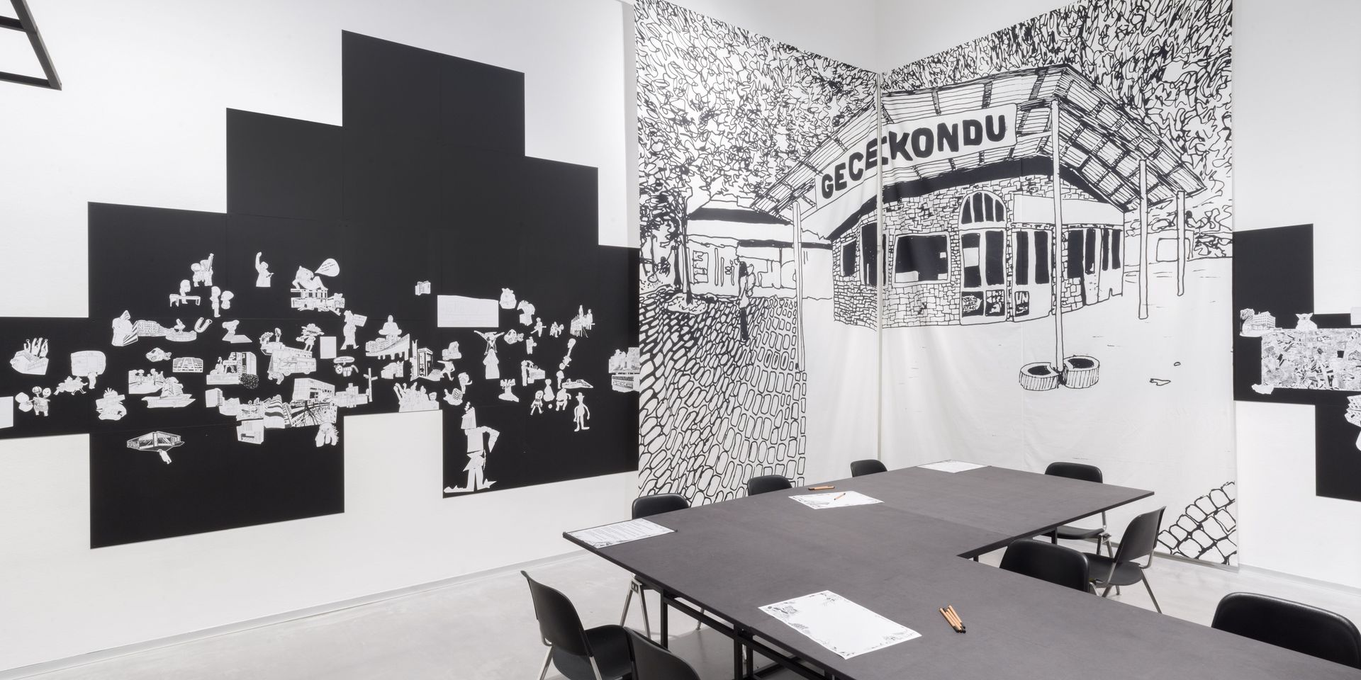 Foto: Blick in den Ausstellungsraum von „Kotti-Shop/SuperFuture“ mit einem großen schwarzen Tisch, Stühlen und Collagen in schwarz-weiß an den Wänden.
